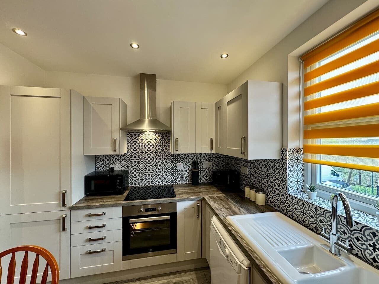 Una cocina moderna con armarios blancos, una campana extractora de acero inoxidable, horno empotrado, microondas y una ventana con persianas amarillas.