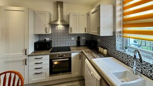 A modern kitchen with white cabinets, a stainless steel range hood, a built-in oven, a microwave, and a window with yellow blinds. - Waterloo House – Contractors, Families, Holidays (Nottingham)