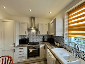 Una cocina moderna con armarios blancos, una campana extractora de acero inoxidable, horno empotrado, microondas y una ventana con persianas amarillas.