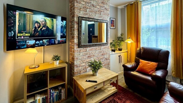 A living room with a brick wall, a mounted TV, a leather armchair, a small table, and a bookshelf. - Waterloo House – Contractors, Families, Holidays (Nottingham)