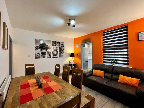 A dining area with a wooden table and chairs, a black leather sofa with orange cushions, and a wall with framed pictures and a sign.
