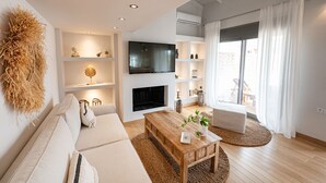 Ein modernes Wohnzimmer mit einer Couch, Couchtisch und Fernseher. Regale mit Dekorationselementen und ein Kamin sind ebenfalls vorhanden.