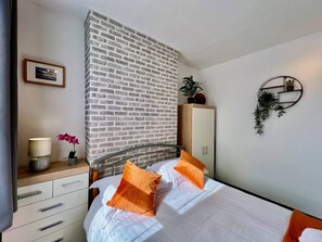 Ett hotellrum med en accentvägg i tegelmönster, en säng med vita sängkläder och orange kuddar, ett sängbord med en lampa och en vas med blommor, en garderob och en vägghylla med växter.