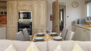 A modern kitchen with wooden cabinets, a built-in oven, and a dining area with a table set for two. - Hazel Lodge (Newton Abbot)