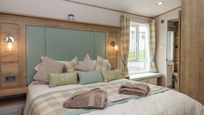 A bedroom with a bed, pillows, a bedside table, a window with curtains, and a wooden wardrobe. - Hazel Lodge (Newton Abbot)