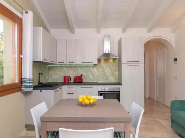 Une cuisine moderne avec des armoires blanches, un comptoir noir et une hotte de cuisinière en acier inoxydable. On y voit une fenêtre avec un rideau, un bol de citrons sur la table et un canapé vert à l’arrière-plan.