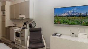 Une cuisine compacte dotée d’appareils électroménagers modernes, un coin repas avec une chaise, et un téléviseur fixé au mur qui diffuse un paysage de montagne.