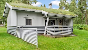 A small, white cabin with a sloped roof covered in grass, a wooden deck, and a metal fence.