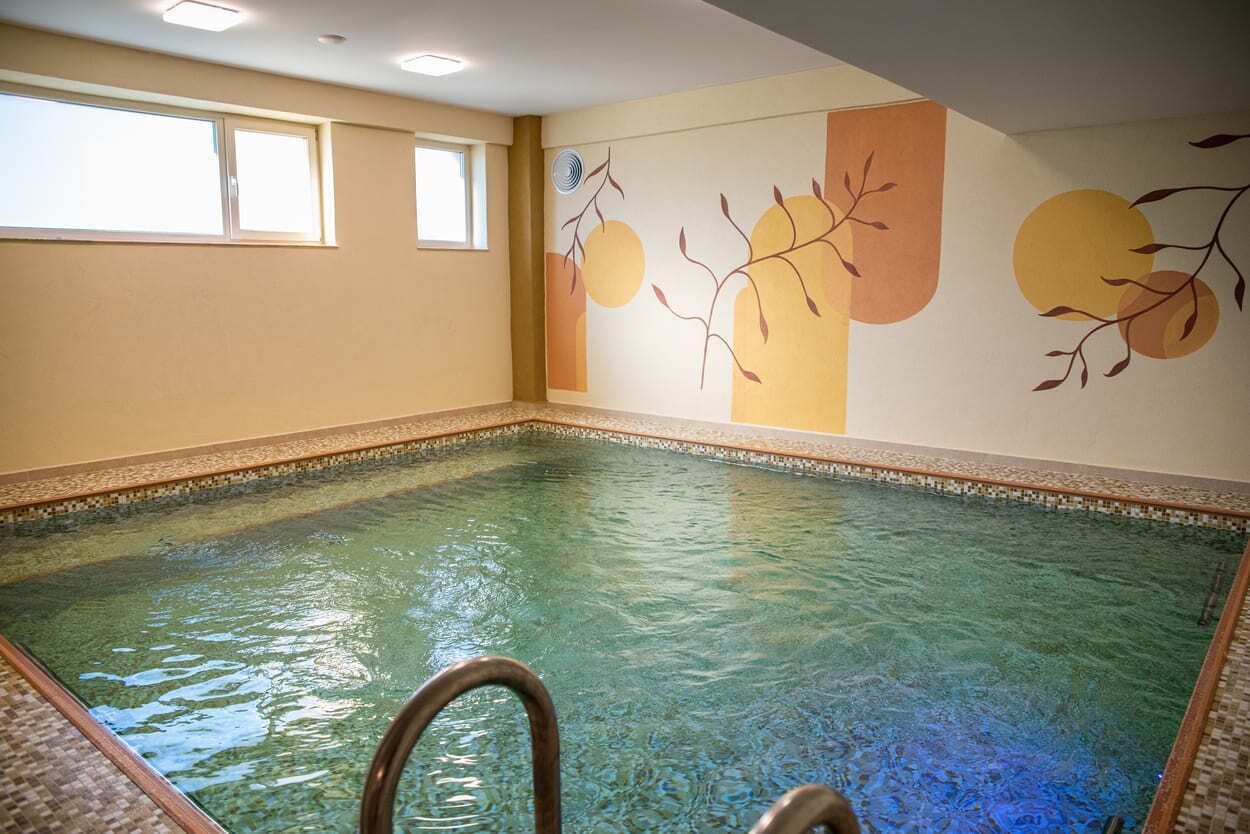 En indendørs swimmingpool med klart vand, flisekant og et vægmaleri med abstrakte designs.