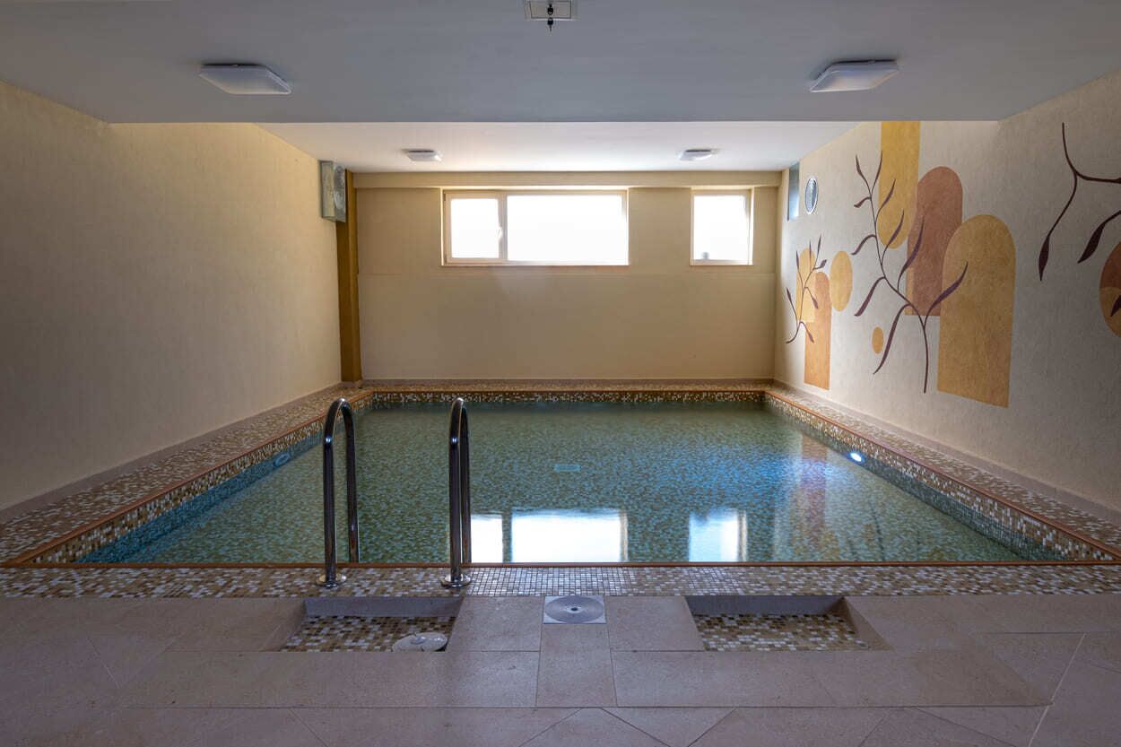 Una piscina interior con agua cristalina, piso de baldosas y un mural en la pared.