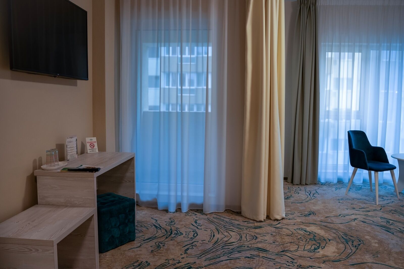 A hotel room with a desk, a chair, a TV mounted on the wall, and a window with sheer curtains.