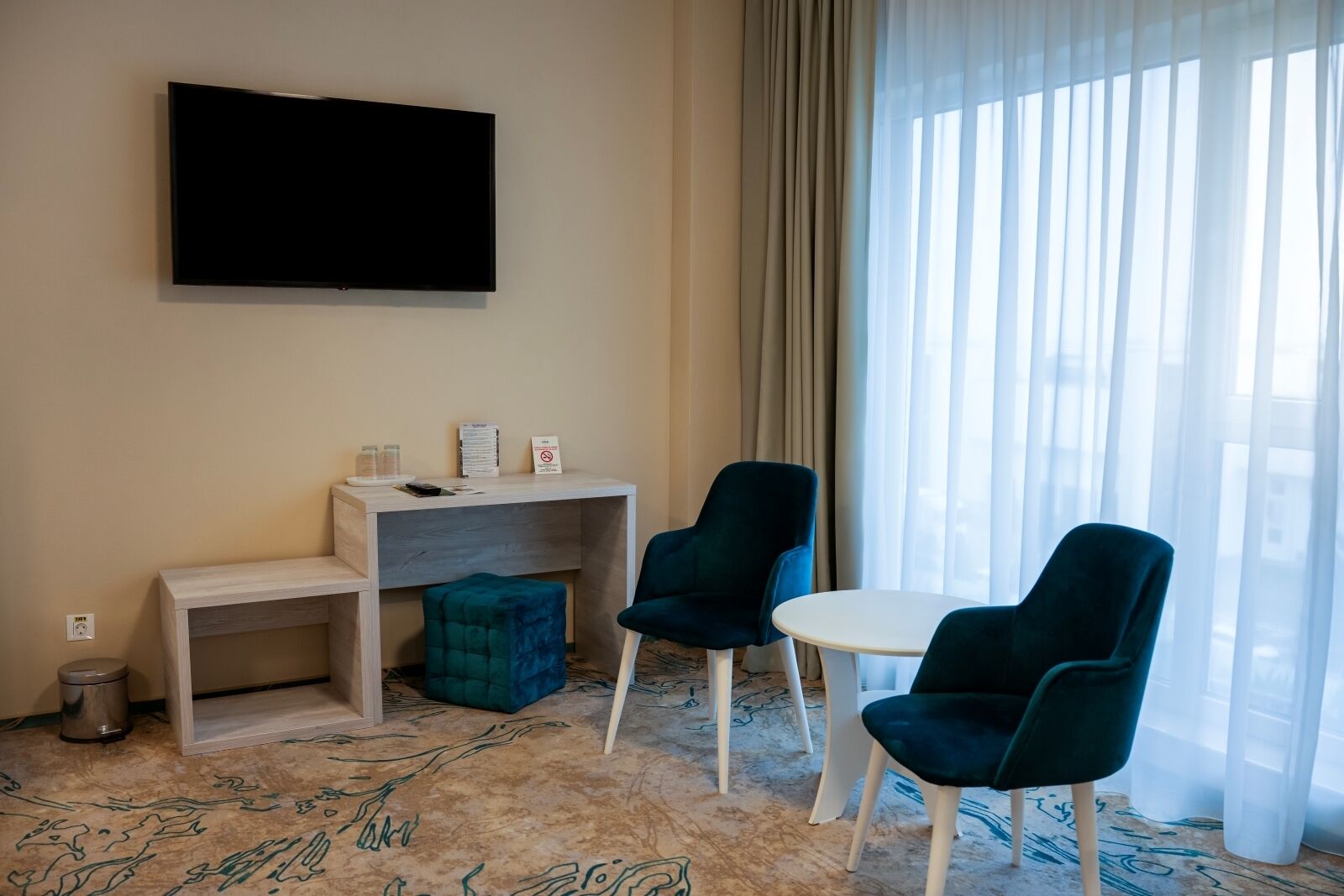 A hotel room with a flat-screen TV mounted on the wall, a desk with a chair, a small ottoman, and a round table with two chairs.