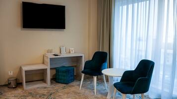 A hotel room with a flat-screen TV mounted on the wall, a desk with a chair, a small ottoman, and a round table with two chairs.
