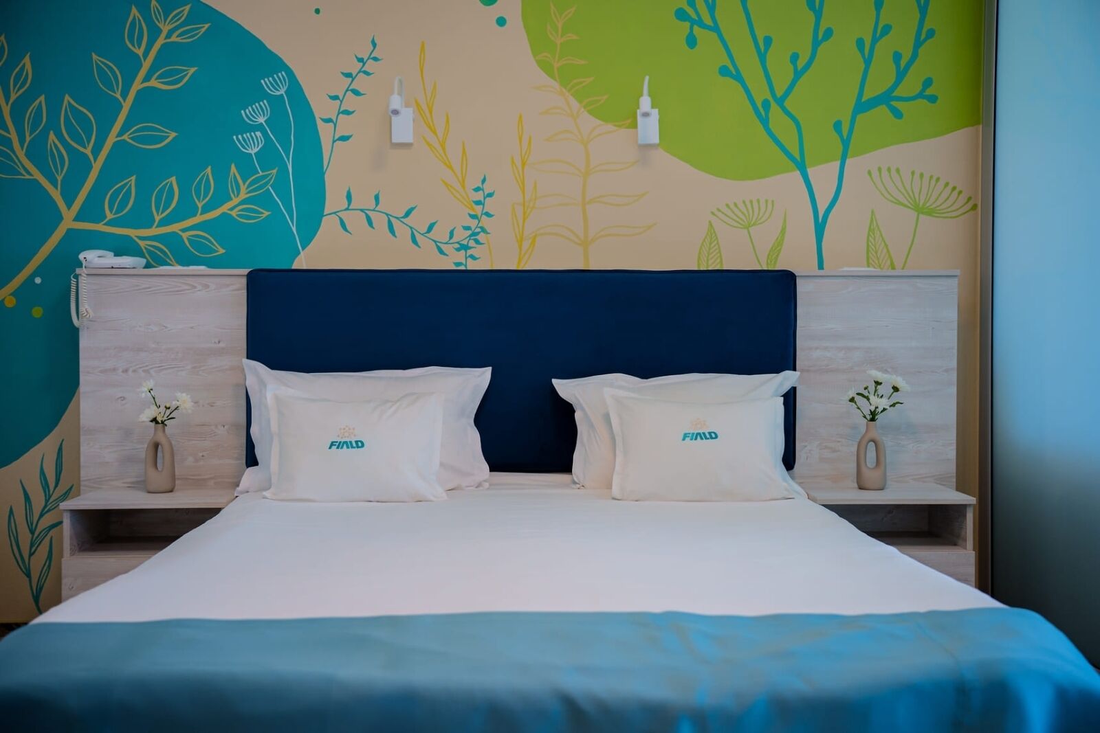 A double bed with two pillows, a dark blue headboard, and a wall with abstract plant designs.