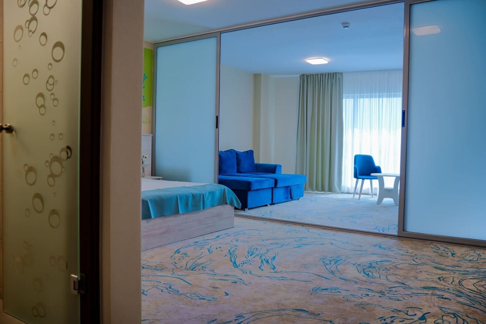 Una habitación de hotel con una cama, un sofá azul, una silla y un amplio ventanal corredizo.