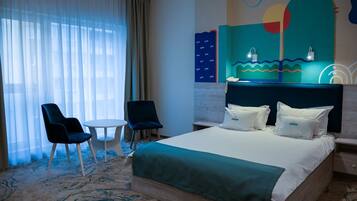 A hotel room with a bed, two chairs, a small table, a nightstand, and a wall with abstract art.
