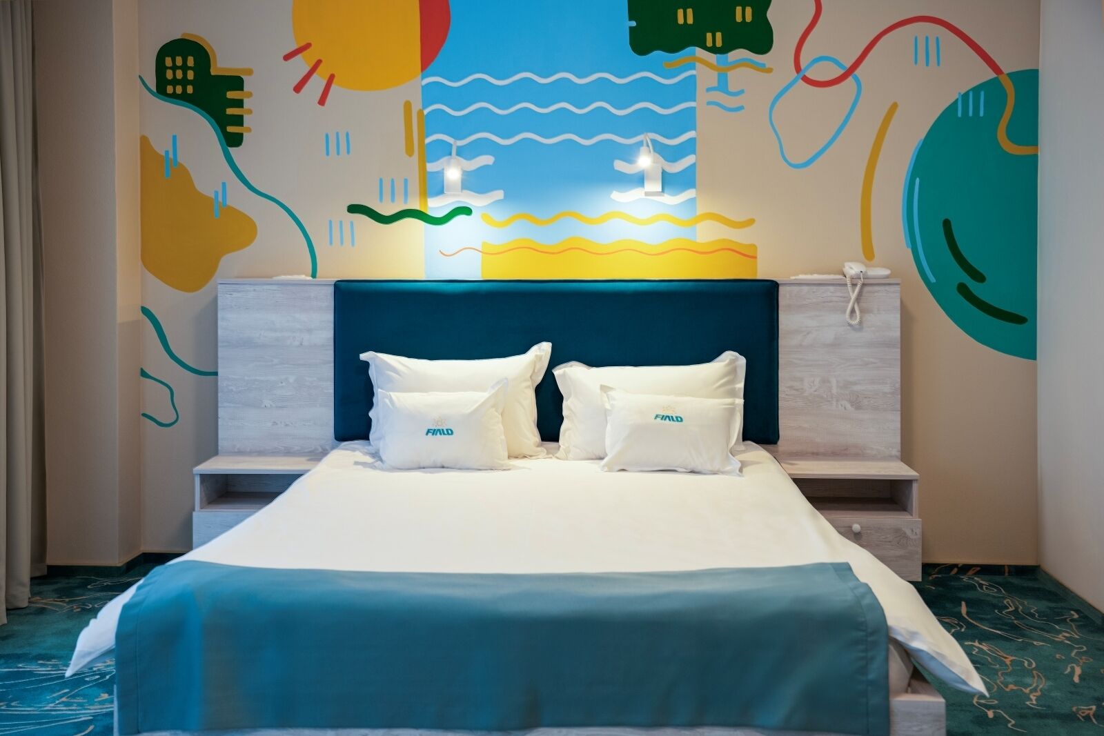 Ein Hotelzimmer mit einem Bett, zwei Kissen, einem Kopfteil, einem Nachttisch und einer Wandmalerei.