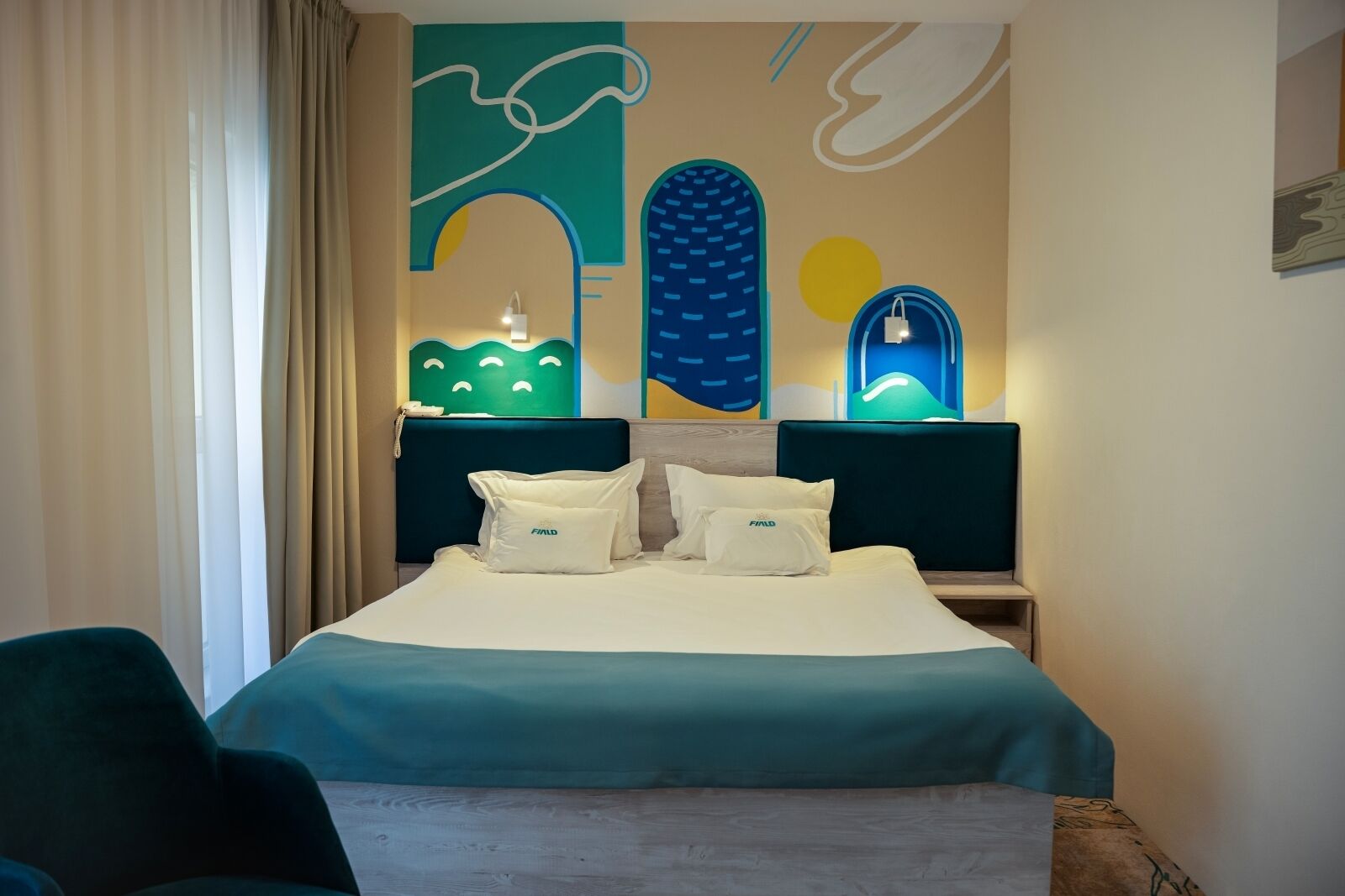 Ein Hotelzimmer mit Bett, zwei Nachttischen, einem Sessel und einer Wand mit abstrakter Kunst.