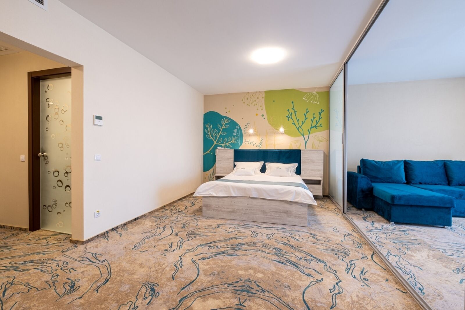 Un dormitorio con cama, un sofá azul, una pared con motivos de hojas y un suelo estampado.