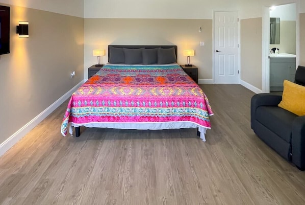 A bedroom with a large bed, two bedside tables with lamps, a dark sofa, and a door to a bathroom.