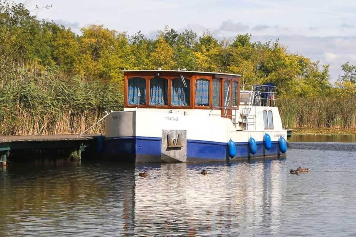 A stationary boat with a cabin, docked at a calm water body surrounded by dense vegetation.