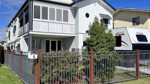 En tvåvånings bostadsbyggnad med en balkong, en inhägnad trädgård och en vit husbil parkerad utanför.