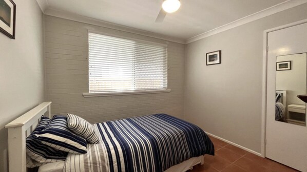A small bedroom with a single bed, a window with blinds, a door with a mirror, and a white ceiling.