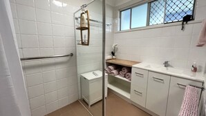 Une salle de bain carrelée de blanc, avec une douche en verre, une étagère en bois où sont posées des serviettes et une fenêtre offrant une vue agréable.