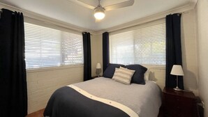 Une chambre à coucher avec un lit, deux lampes de chevet, un ventilateur de plafond et des fenêtres équipées de stores.