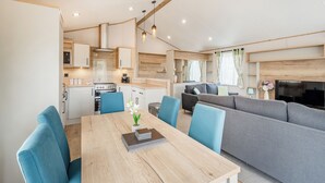 A modern kitchen with built-in appliances, a dining area with a wooden table and blue chairs, and a living area with a grey sofa and TV. - Hot Tub Lodge 3 Pet Friendly (Kirkcudbright)