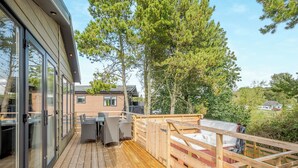 Una terraza de madera con mobiliario exterior, una puerta de cristal y vista a una casa al fondo.