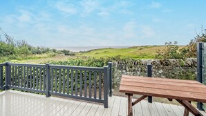 Um deck de madeira com um banco e um muro de pedra, com vista para uma paisagem gramada e o mar.
