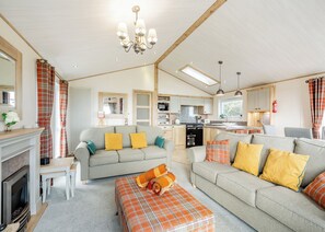A modern living room with a fireplace, sofa, ottoman, and kitchen area with a dining table. - Country and modern cladded lodge with sea views. (Kirkcudbright)