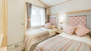 A bedroom with two beds, a wardrobe, a window with curtains, and a headboard with a light fixture. - Country and modern cladded lodge with sea views. (Kirkcudbright)
