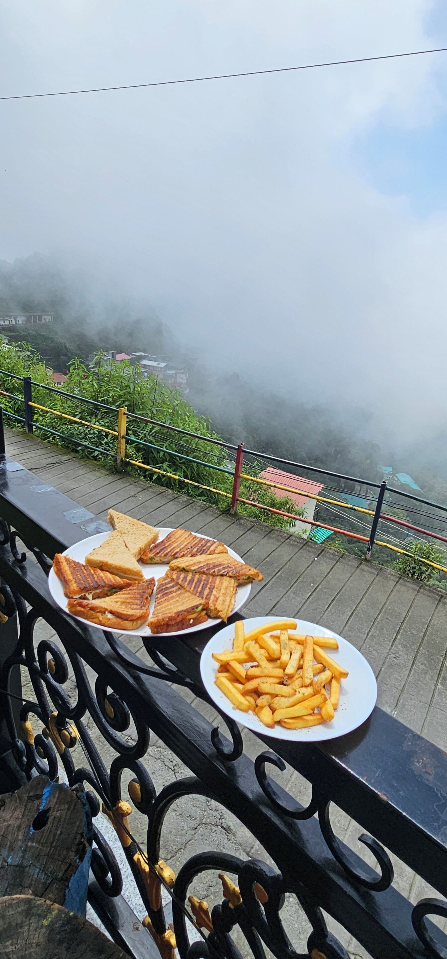 A dining table with a plate of grilled sandwiches and fries, overlooking a misty landscape with buildings in the distance.