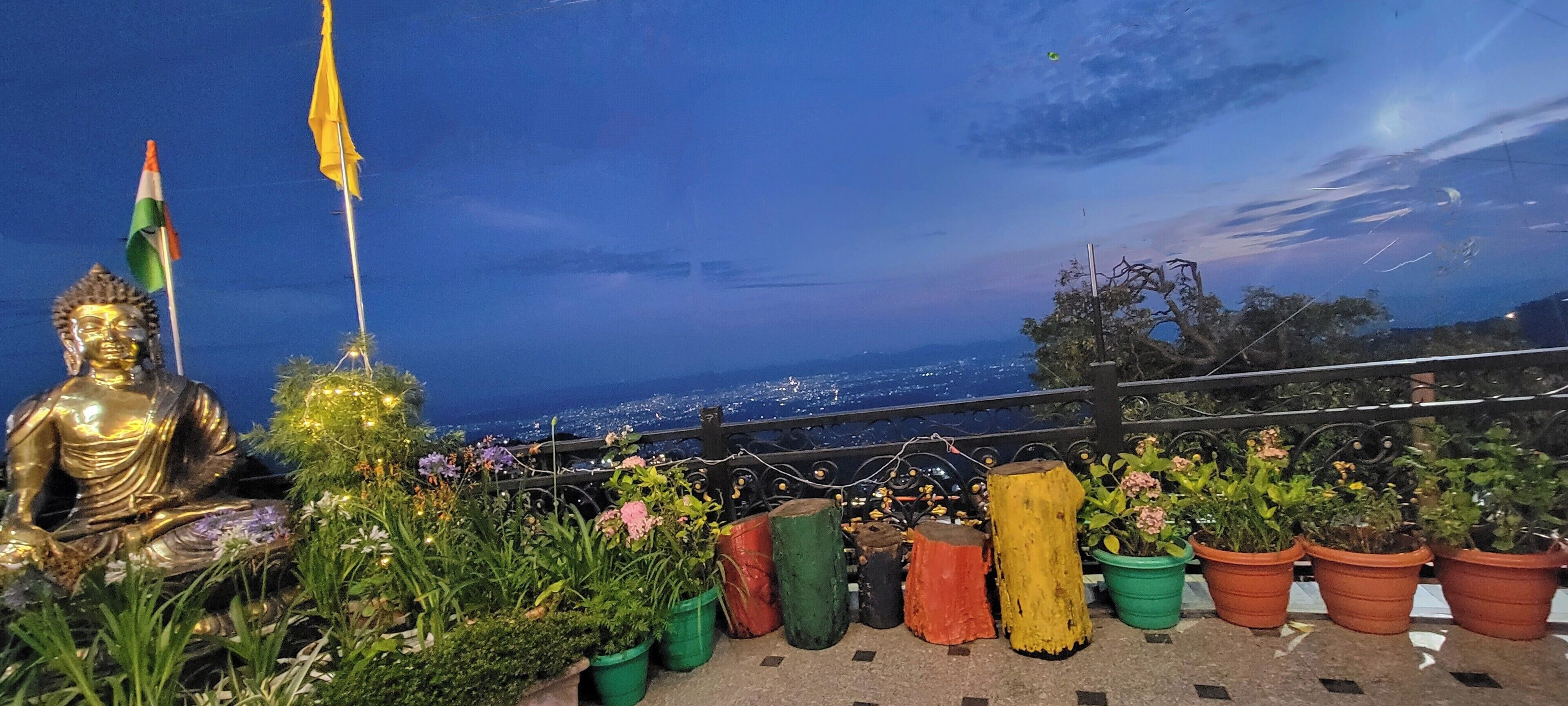 A Buddha statue, flags, and potted plants on a balcony with a city view.