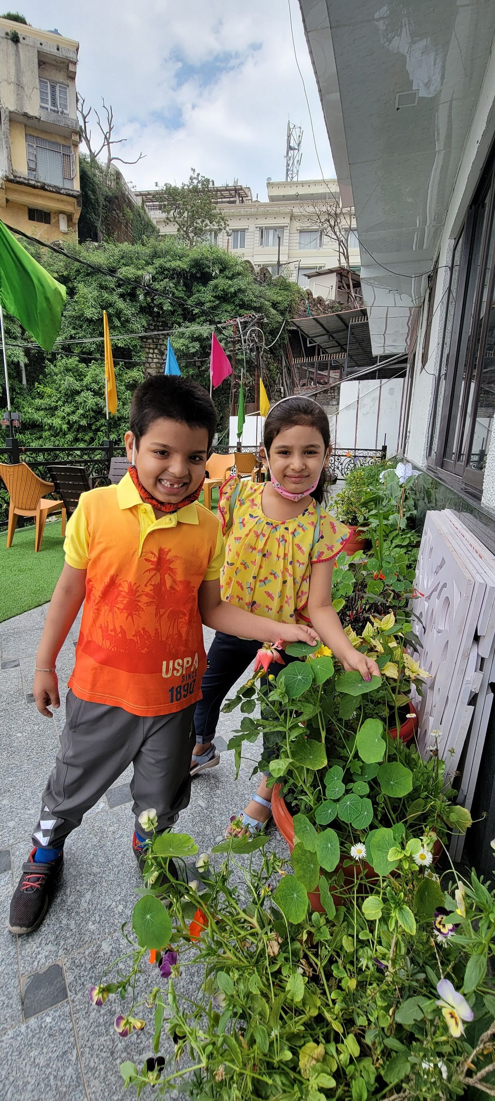 Two children standing next to a potted plant with a variety of flowers.