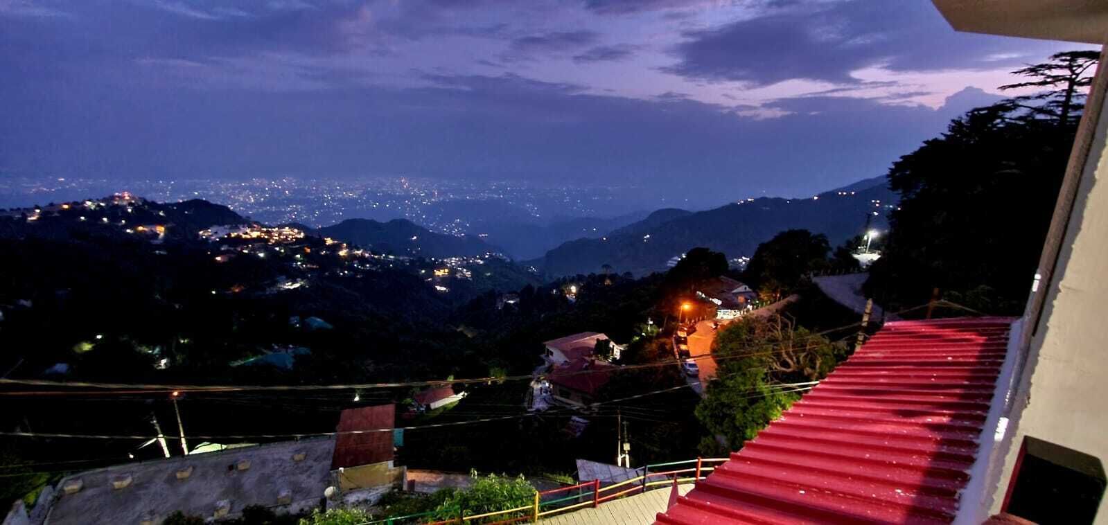 A nighttime view of a mountainous area with scattered lights, a red-roofed building in the foreground, and a distant cityscape.