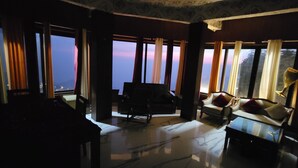 A dimly lit room with large windows, a sofa, and a table. - Casa Arhan Luxury Mussoorie Homestay (Dehradun)