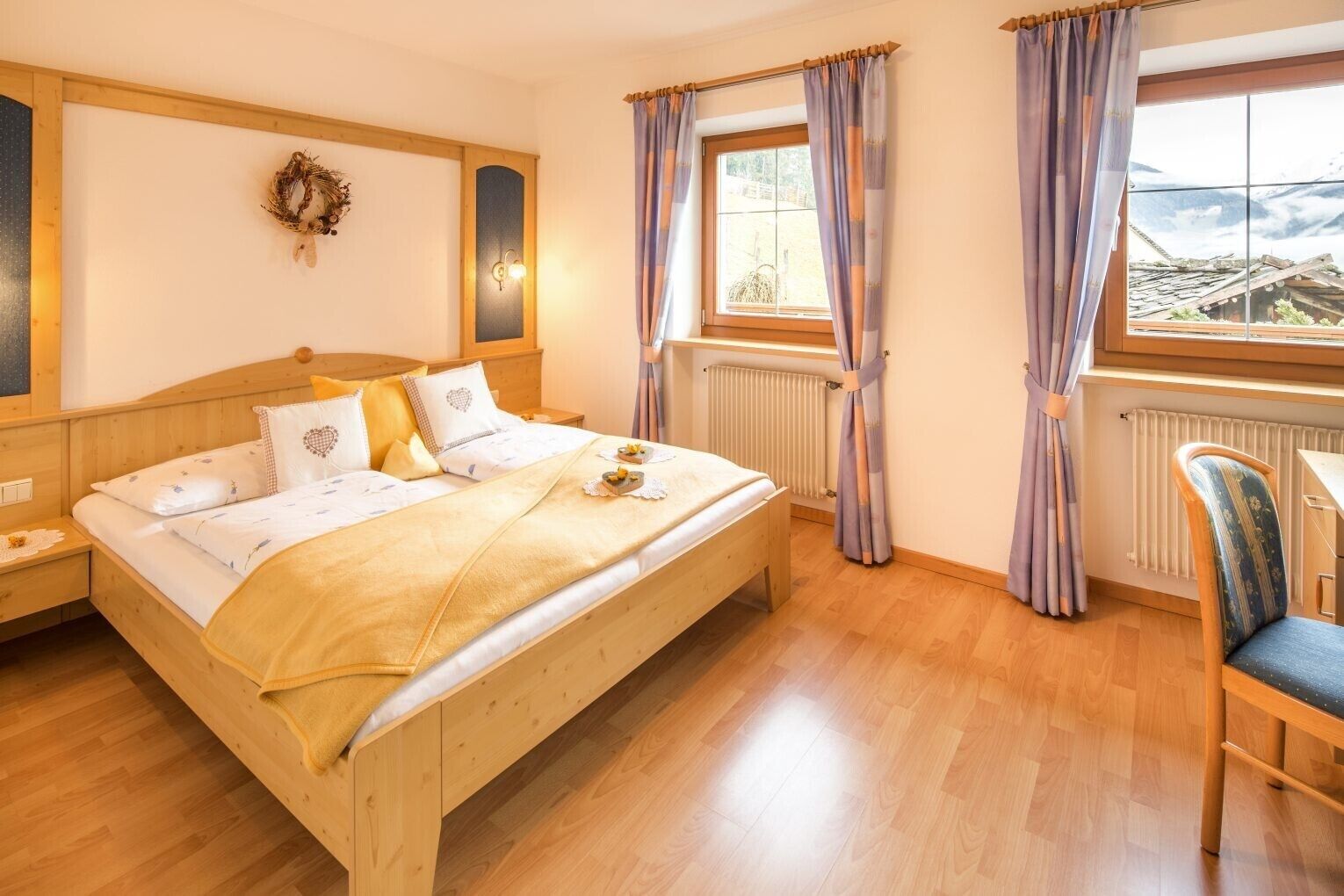 Une pièce comprenant un lit en bois, une table de chevet, une chaise, une fenêtre avec des rideaux et un luminaire fixé au mur.
