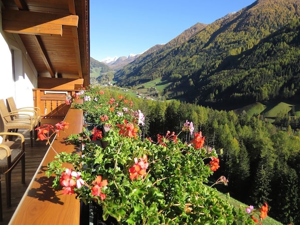 A wooden balcony with wicker chairs and a flower arrangement, overlooking a valley with mountains and a clear sky.
