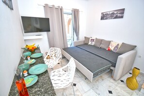 A compact living space with a sofa, a flat-screen TV, a dining area with chairs and plates, and a window with curtains.