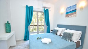 A bedroom with a bed, curtains, a wall-mounted air conditioner, a shelf with a decorative item, and a beach-themed wall art. - Doliva Erietta Apartment with Private Pool, Mountain View, Wi-Fi & Air Conditioning (Corfu)