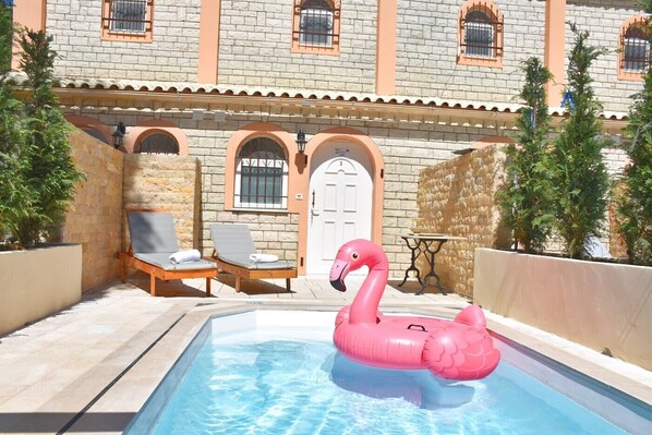 Une piscine avec un flotteur en forme de flamant rose, des chaises longues et un bâtiment doté de fenêtres en arc de cercle et d’une porte.