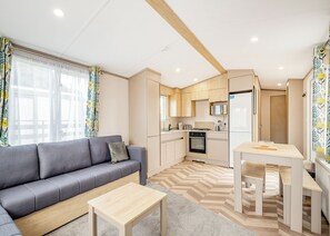 A modern apartment with a grey sofa, a small wooden table, a kitchen with a microwave, stove, and refrigerator, and a patterned curtain.