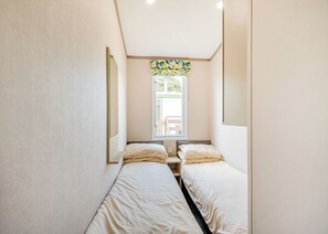 A small, narrow room with two single beds, a window with a view of the outside, and a mirror on the left wall.