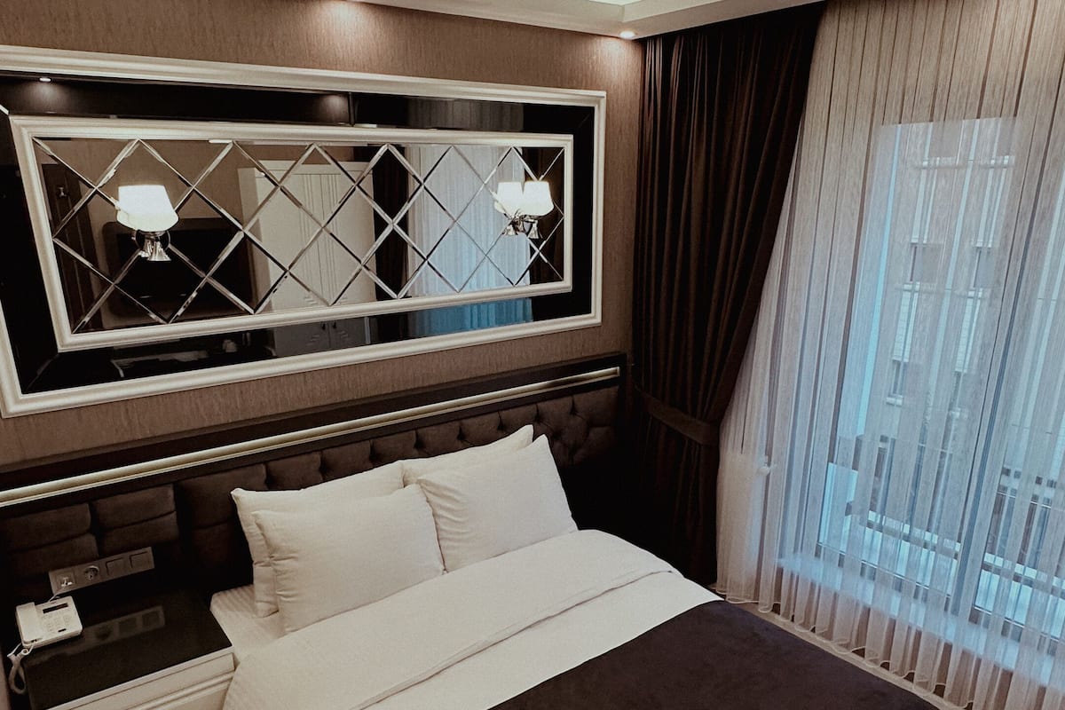 A modern hotel room with a large bed, a nightstand, a mirror, and a view of the city through the window.