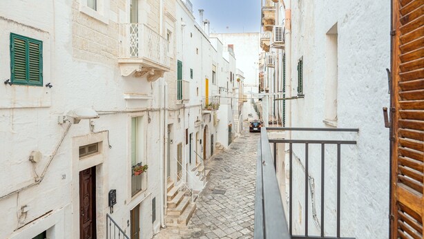 Une ruelle étroite pavée, bordée de bâtiments blancs dotés de balcons et de volets verts.