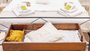 Un lit bien fait, avec des serviettes pliées, et une commode en bois contenant un coussin jaune et un coussin blanc.