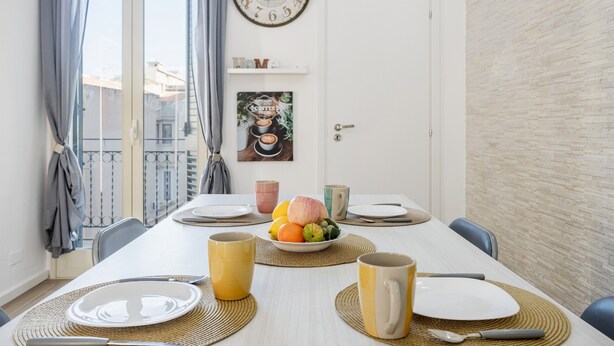 A dining area with a table set for four, a clock, and a view of the outside.